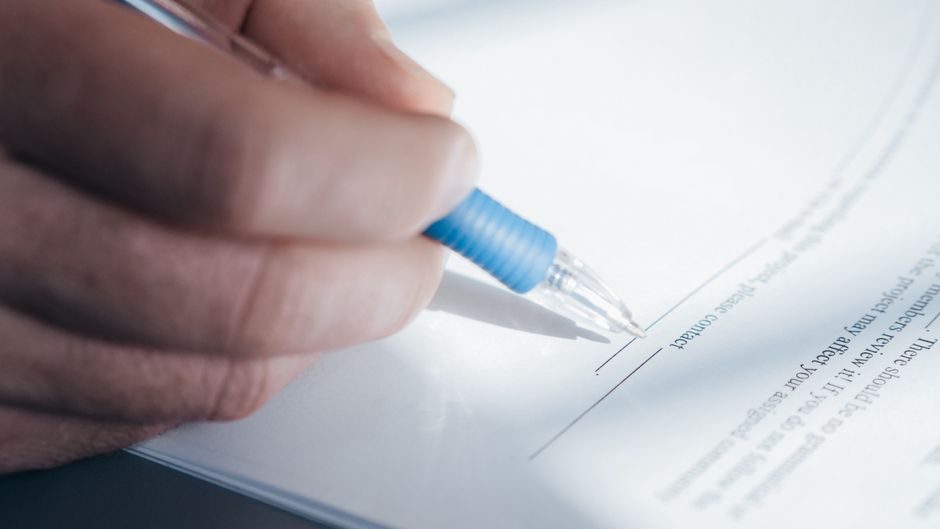 A close-up image showing a hand holding a pen while signing a document.