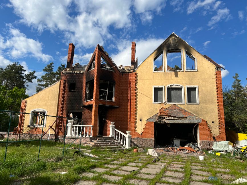 A burnt brick house stands in ruins with clear skies and greenery surrounding it.